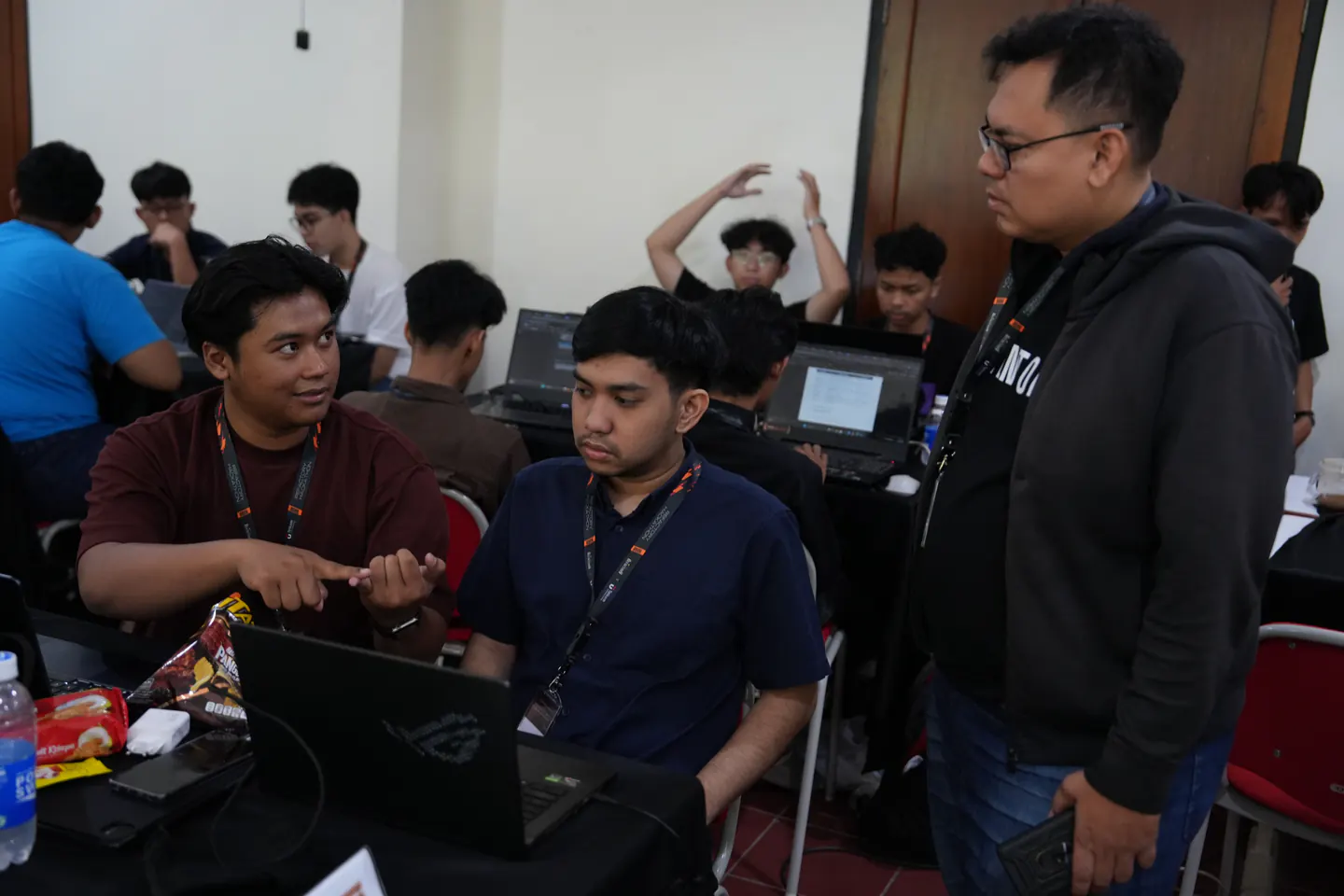 Wildan gestures while discussing work with a teammate at a laptop during the Refactory Hackathon.